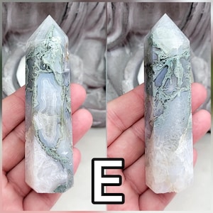 May include: A polished crystal tower featuring a hexagonal point and a smooth, multifaceted surface. The stone displays a blend of translucent white, soft gray, and mossy green dendritic inclusions, with the letter E displayed in the center.