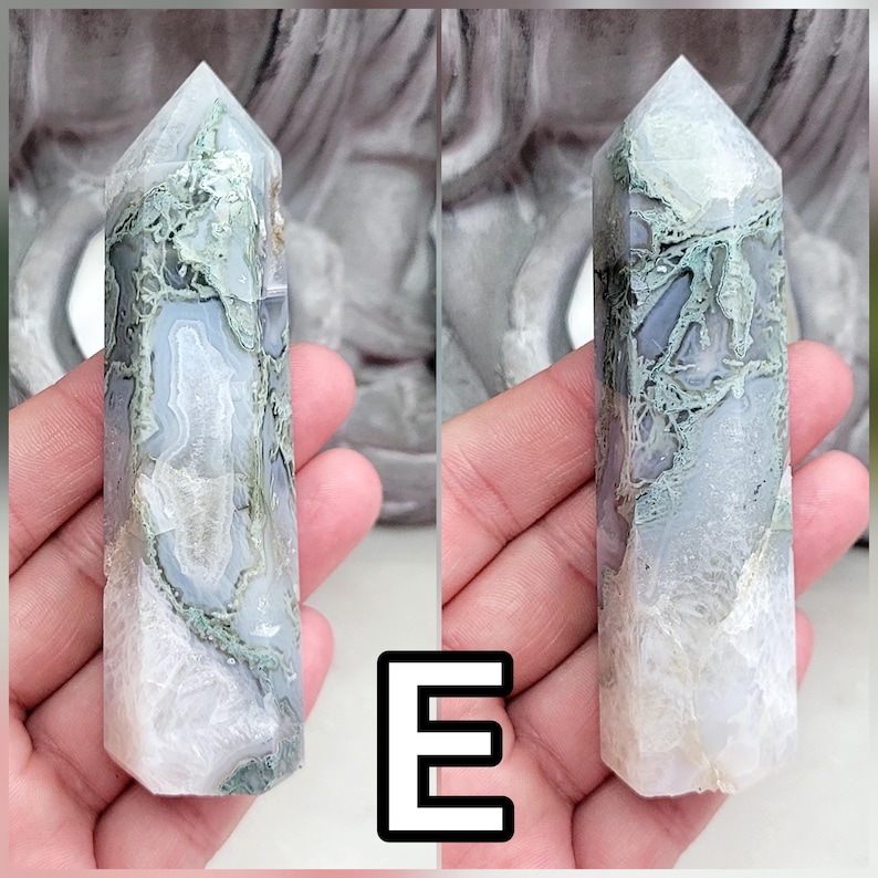 May include: A polished crystal tower featuring a hexagonal point and a smooth, multifaceted surface. The stone displays a blend of translucent white, soft gray, and mossy green dendritic inclusions, with the letter E displayed in the center.
