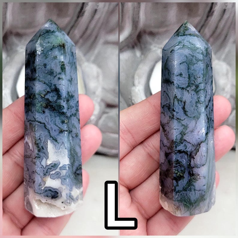 May include: A polished moss agate crystal tower featuring a hexagonal point and a smooth, cylindrical body. The stone displays a translucent light blue and white base with intricate, dark green, branch-like dendritic inclusions throughout.