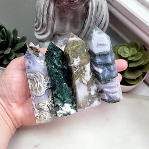 May include: Four polished crystal towers featuring distinct mineral patterns. The first is lavender with white inclusions, the second is deep forest green, the third is white with gold-toned mossy veins, and the fourth is blue-grey with banded layers.