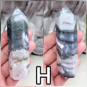May include: A polished crystal tower featuring a hexagonal point, displaying layers of translucent lavender and white stone with dark green moss-like inclusions throughout the structure. The letter H is displayed in a white box at the bottom center.