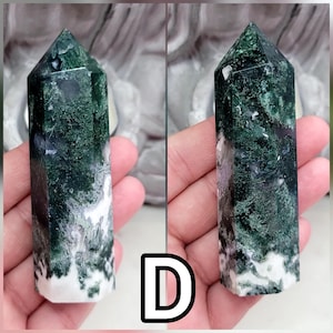 May include: A polished moss agate crystal tower featuring a hexagonal prism shape with a pointed top. The stone displays deep forest green moss-like inclusions, translucent areas, and opaque white mineral patterns throughout the entire structure.