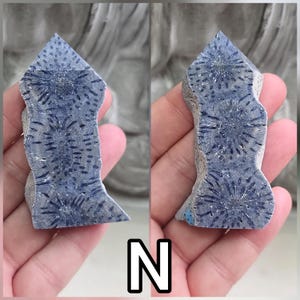 May include: A polished, tower-shaped specimen of blue fossilized coral featuring distinct, dark blue star-shaped patterns throughout its surface. The stone displays a natural, textured edge on one side and a pointed top, showcasing organic radial structures.