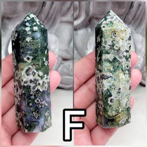 May include: A polished moss agate crystal tower featuring a hexagonal point. The stone displays a complex pattern of deep forest green, translucent gray, and creamy white inclusions, with moss-like dendritic formations throughout the mineral structure.
