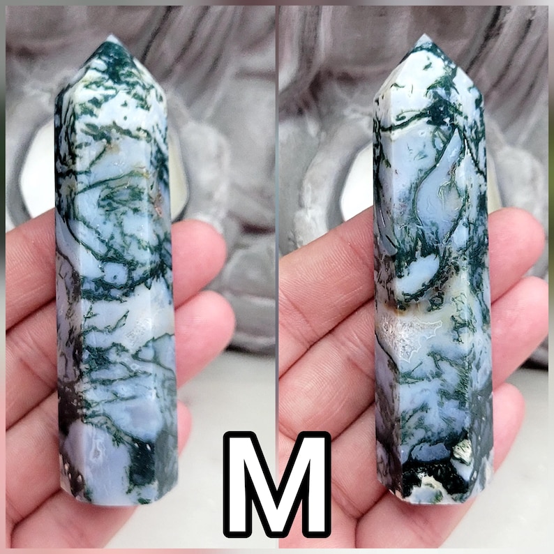 May include: A polished moss agate crystal tower with a hexagonal shape, featuring a translucent white base with intricate, dark green, branch-like inclusions throughout the stone. The letter M is displayed in a black and white square below.