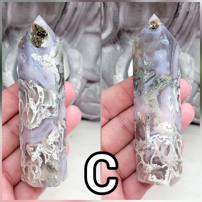 May include: A polished crystal tower with a hexagonal point, featuring a translucent lavender and white base with intricate, moss-like green and brown inclusions throughout the stone. The top of the crystal displays a small, metallic gold cluster.