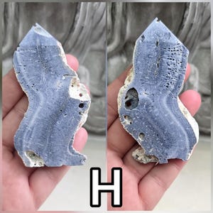 May include: A polished, irregular slab of blue-grey mineral specimen featuring a distinct radial, fibrous crystalline structure and natural porous inclusions. The specimen displays a pointed top and a curved, organic shape with a rough, light-colored edge.