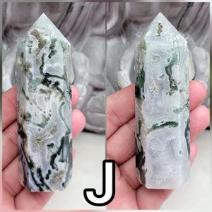May include: A polished moss agate crystal tower featuring a hexagonal prism shape with a pointed top. The stone displays a translucent white and grey base with intricate, branching dark green inclusions that resemble moss-like patterns throughout.
