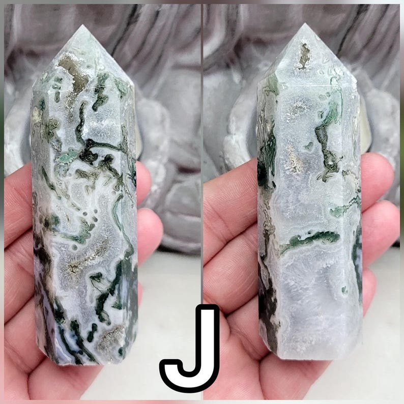 May include: A polished moss agate crystal tower featuring a hexagonal prism shape with a pointed top. The stone displays a translucent white and grey base with intricate, branching dark green inclusions that resemble moss-like patterns throughout.