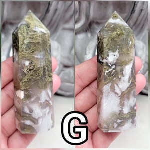 May include: A polished moss agate crystal tower featuring a hexagonal point and a smooth, multifaceted surface. The stone displays a translucent, cloudy white base with intricate, moss-like inclusions in shades of olive green and earthy brown.