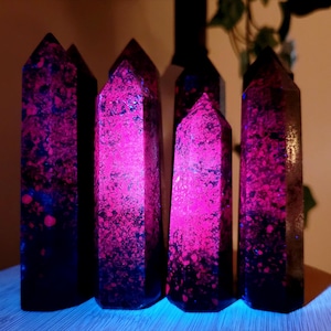May include: A collection of six dark, elongated crystal obelisks. The crystals have a speckled pattern of bright pink and blue against a dark background. The crystals are standing upright, with a light source illuminating them from below.