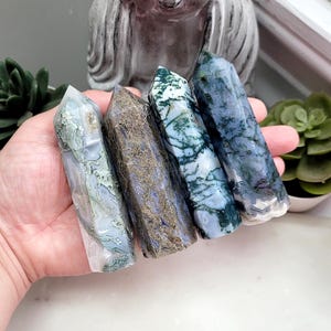 May include: Four polished crystal towers held in a palm. The first is translucent with green moss-like inclusions. The second is a dark, textured brown and blue. The third is white with deep green dendritic patterns. The fourth is a blue-grey stone.