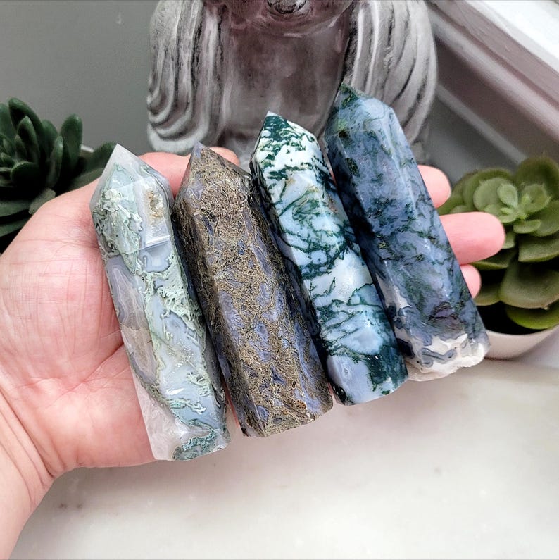 May include: Four polished crystal towers held in a palm. The first is translucent with green moss-like inclusions. The second is a dark, textured brown and blue. The third is white with deep green dendritic patterns. The fourth is a blue-grey stone.