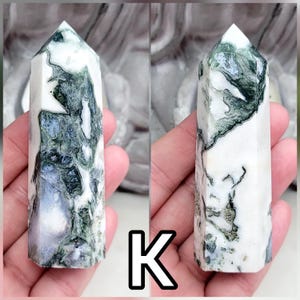 May include: A polished stone tower with a hexagonal base and a pointed top, featuring a white base color with intricate, moss-like dark green inclusions and translucent patches throughout the crystalline structure, marked with the letter K.