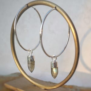 May include: Silver hoop earrings with labradorite pendants. The earrings are displayed on a gold-coloured metal stand. The labradorite pendants have a silver cap and a shimmering, iridescent appearance.