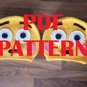 May include: Two yellow crocheted emoji hats. Each hat has white eyes with black pupils, black eyebrows, and a pink tongue. The image has the words "PDF PATTERN" in red across the hats.