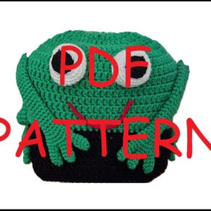 May include: A green crocheted frog hat with large white and black eyes. The hat has a black base and the text "PDF PATTERN" in red. The hat is handmade and suitable for a costume or novelty item.