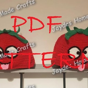 May include: Two crocheted tomato hats with googly eyes and a tongue sticking out. The hats are red with green leaves and are sitting on a shelf. The text "PDF PATTERN" is written in red letters on the image.
