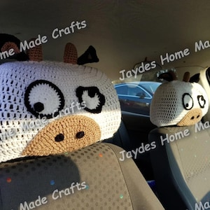 May include: Two crocheted cow headrest covers for car seats. The headrests are white with brown accents for the nose, ears, and eyes. The eyes are black with white pupils.