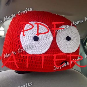 May include: A red, crocheted headrest cover with large white eyes and black pupils. The word "PATTERN" is written across the front in red. The headrest is installed in a car.