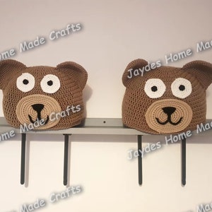 May include: Two brown crocheted bear hats with white eyes and a brown nose. The hats are displayed on a shelf with black metal brackets.