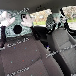 May include: Two crocheted headrest covers in a car. The headrest covers are light blue with black eyes, a black nose, and white hands. The text "Jaydes Home Made Crafts" is visible in the image.