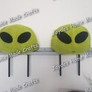 May include: Two green alien head-shaped crocheted ornaments with black eyes, mounted on a shelf with black metal brackets.