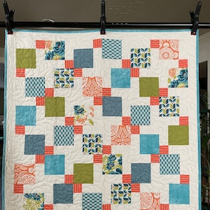 May include: A white quilt with a blue, green, and orange patchwork design. The quilt is made up of squares with different patterns and colors. The quilt is hanging on a black rod.