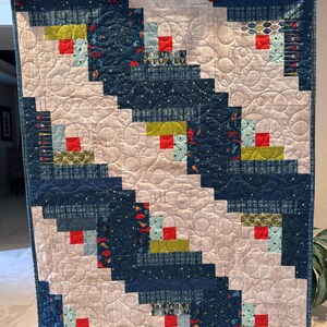 May include: A handmade quilt with a log cabin design. The quilt features a pattern of alternating light beige and dark blue blocks. Each block has a red square center, surrounded by various colorful fabrics. The quilt is hanging from a black bar.