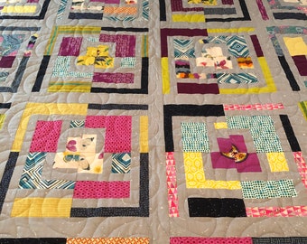 May include: A quilt with a geometric pattern in shades of gray, yellow, pink, and blue. The quilt is made up of squares and rectangles, and the pattern is repeated throughout the quilt.