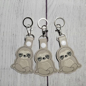 May include: Three beige keychains with embroidered sloth designs. Each keychain features a smiling sloth with closed eyes and a light brown body.
