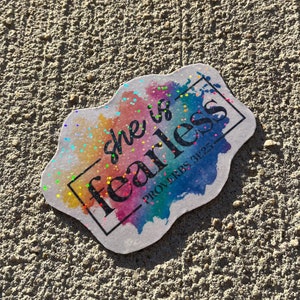 May include: A sticker with the words "she is fearless" in black script, framed by a black rectangle. The background features a watercolor design with rainbow colors and glitter accents. The text "PROVERBS 31:25" is below the main text.