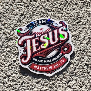 May include: A sticker with a baseball theme, featuring the word "JESUS" in a bold, stylized font. The sticker includes the text "TEAM" and "GO AND MAKE DISCIPLES MATTHEW 28:19" in a red and white color scheme.
