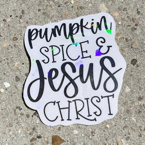 May include: A white sticker with the words "pumpkin SPICE & Jesus CHRIST" in black script. The sticker has a holographic, iridescent sheen. The sticker is on a gray, textured surface.