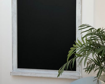 Può includere: Una lavagna rettangolare con una cornice in legno bianco anticato. La lavagna è montata su una parete bianca. Una piccola pianta verde in un vaso bianco e un tavolo azzurro sono in primo piano.