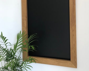 Può includere: Una lavagna nera con cornice in legno, montata su una parete bianca. Una pianta verde è in primo piano.