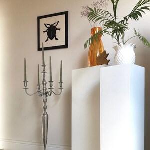 May include: Two white square pedestals, one taller than the other, with a silver candelabra with six candles, a vase with dried flowers, and a white pineapple-shaped vase on top. The pedestals are on a wood floor.