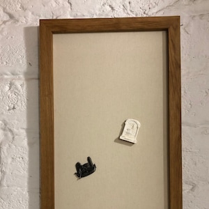 May include: A rectangular wooden framed bulletin board with a beige fabric interior. Two decorative magnets are attached: a black castle and a white tombstone. The board is mounted on a white brick wall.