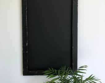 Può includere: Una lavagna nera rettangolare con una cornice in legno nero anticato. La lavagna è montata su una parete bianca, con una pianta verde in primo piano.