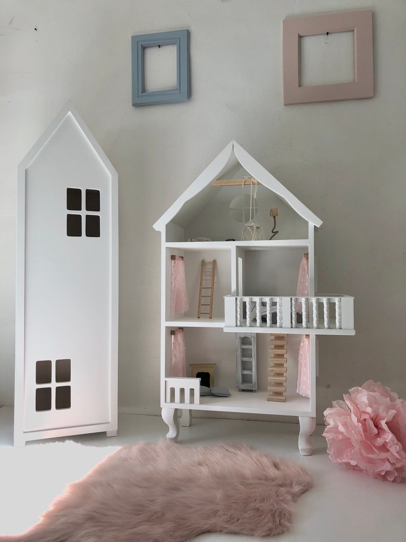 May include: White wooden dollhouse with a pink rug, a pink flower, and two white frames on the wall. The dollhouse has a balcony, a ladder, and a fireplace.