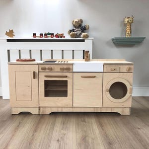Puede incluir: Un juego de cocina de madera con una encimera blanca, una estufa con cuatro quemadores, un horno, un fregadero, un lavavajillas y una lavadora. La cocina está hecha de madera clara y tiene un diseño moderno.