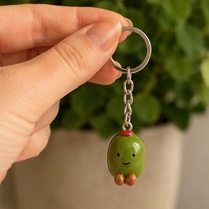 May include: A keyring featuring a smooth, olive-green, oval-shaped charm with a small red cap on top and two tiny brown feet at the bottom. The charm has a simple, hand-painted face with two black dot eyes and a thin, curved line for a smile.