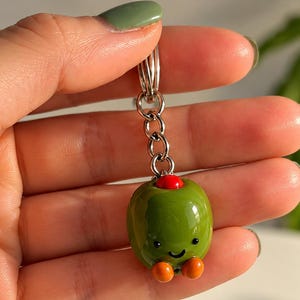 May include: A keychain featuring a smooth, glossy green olive charm with a small red pimento center, a tiny smiling face, and two small orange feet attached to a silver-toned metal chain and a circular split ring for keys.