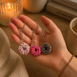 May include: Three miniature doughnut keychains attached to silver-toned metal rings and chains. One doughnut is white with multicoloured sprinkles, one is pink with multicoloured sprinkles, and one is dark brown with white icing drizzle patterns.