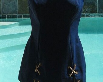 May include: A navy blue one-piece swimsuit with thick straps and a scoop neckline. The hemline features decorative gold-colored rope and anchor details. The swimsuit is displayed on a mannequin in front of a pool.