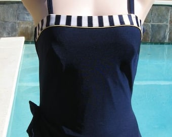 May include: A navy blue swimsuit with a white and black striped band at the top. The swimsuit has a gathered skirt and thin straps.