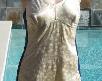 May include: A vintage-style, cream-colored camisole with a subtle polka dot pattern. The top features thin straps, a V-neckline, and navy blue side panels. The garment is displayed on a mannequin near a swimming pool.