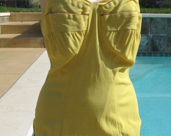 May include: A vintage-style yellow swimsuit with a sweetheart neckline and thin straps. The one-piece suit features decorative pockets and trim. The mannequin is positioned in front of a pool, showcasing the swimwear's design and color.