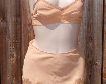 May include: A peach-colored lingerie set, consisting of a bra and shorts. The bra has thin straps and lace trim. The shorts feature floral lace detailing along the hem. The set is displayed on a mannequin against a wooden background.
