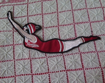 May include: Embroidered patch featuring a swimmer in a red swimsuit and cap with white stripes. The patch has the word "Jantzen" in a script font. The patch is outlined in black.